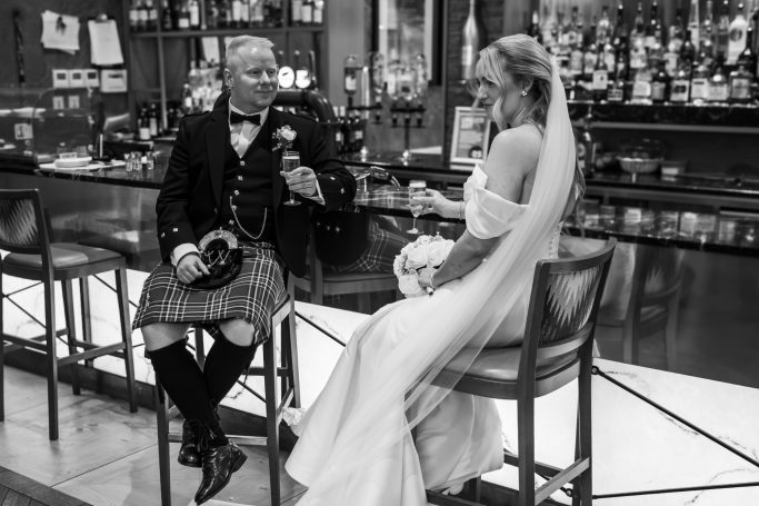 A wedding couple sitting at a bar, the groom in a kilt and the bride in a white gown.