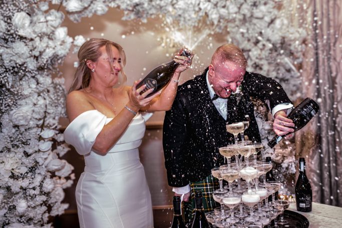 Bride and groom joyfully spraying champagne over a pyramid of glasses at their wedding.