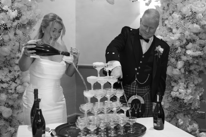 Bride and groom pouring champagne into a tower of glasses at a wedding celebration.