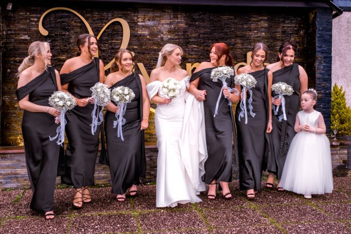 Bride in a white dress stands with bridesmaids in black dresses, holding bouquet arrangements.