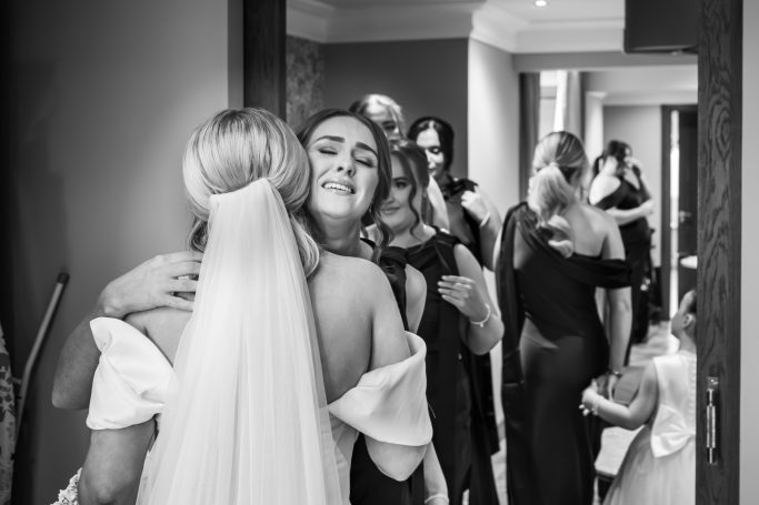 A bride embraces a woman in a wedding setting, with guests in the background.