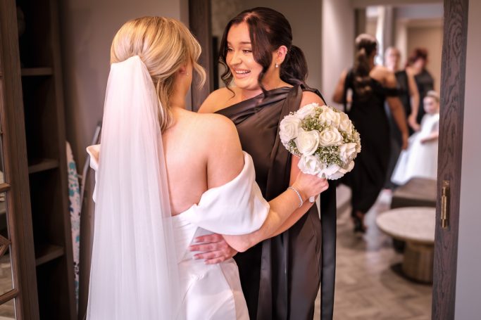 Two women embrace, one in a wedding dress and the other in a black gown, smiling.