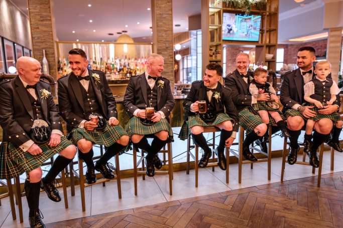 A group of men in kilts seated at a bar, smiling and chatting together.