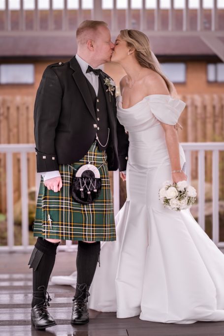 Bride and groom share a kiss, with the groom in a kilt and the bride in a white gown.