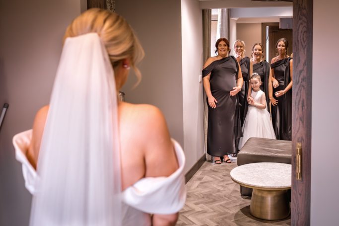 A bride in white attire stands in a doorway, facing friends and family in elegant outfits.