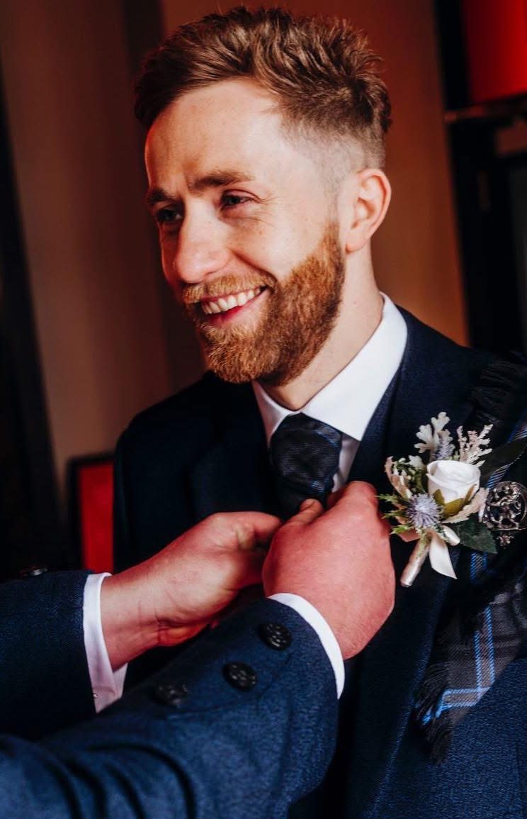 Neill McGlynn in a suit adjusting his tie, with a flower pinned to his lapel captured by Stephen McCluskey Photography