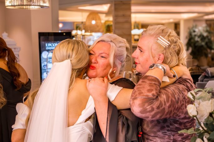 Three women embrace warmly at a social event, with one in a wedding dress.