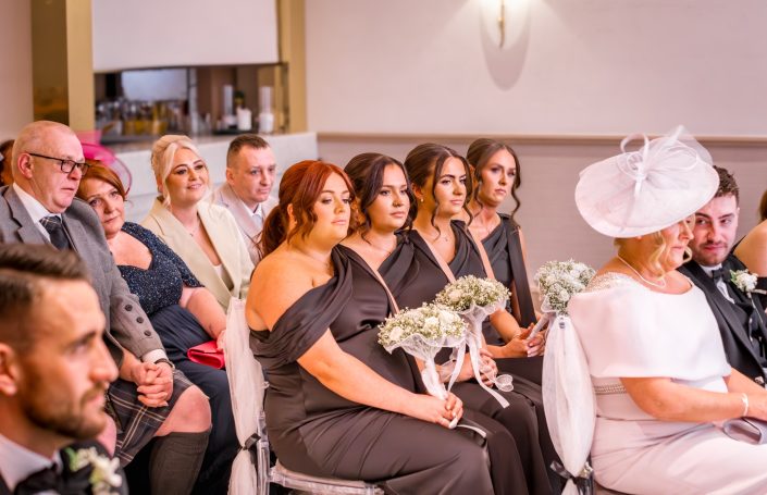 Guests seated at a wedding, dressed in formal attire, with floral arrangements in hand.