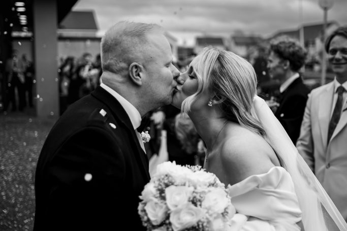 Bride and groom share a kiss, surrounded by wedding guests, in a festive outdoor setting.