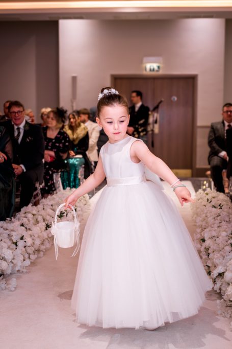 A young girl in a white dress carries a small bag while walking down an aisle.