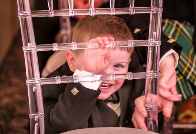 A joyful child in formal attire peeks through a transparent chair back.