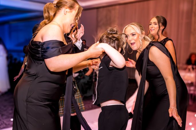 Three women in elegant black outfits dancing, assisting a child on the dance floor.